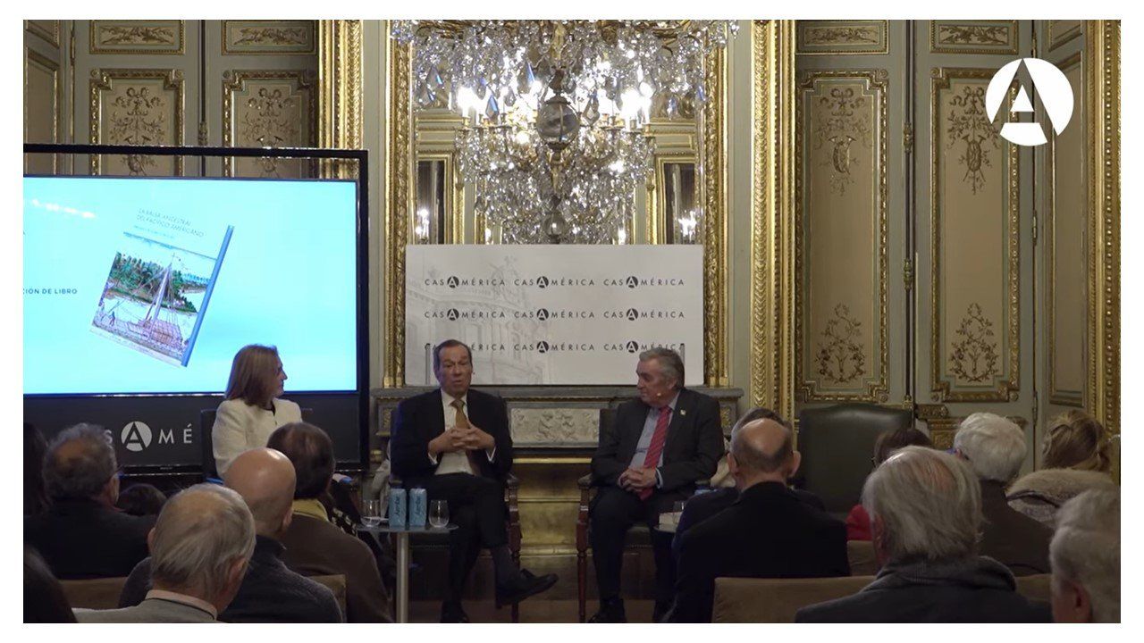 Conversatorio en un salón palaciego de Casa de América con la portada del libro proyectada en pantalla