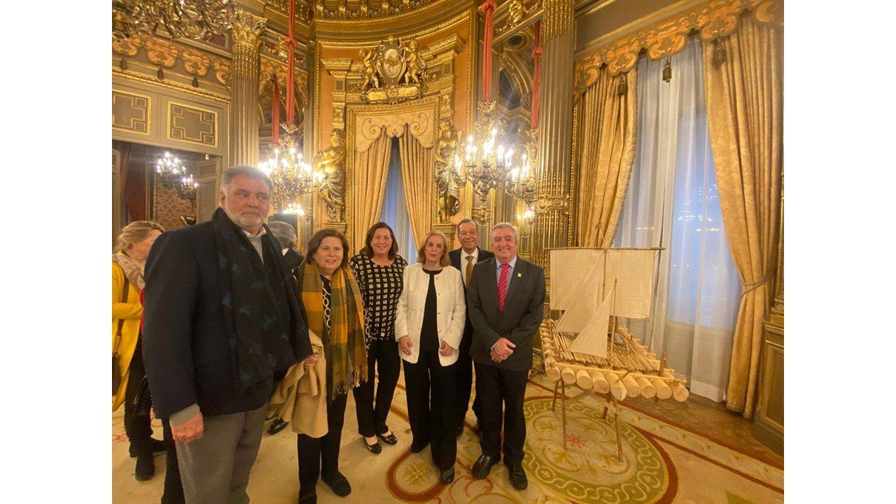 Grupo de asistentes junto a la maqueta de la balsa en un salón con candelabros del Palacio de Linares